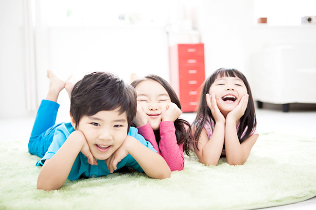 남자 아동 1명과 여자 아동 2명이 바닥에 엎드려 얼굴을 손에 기대고 웃고 있는 이미지