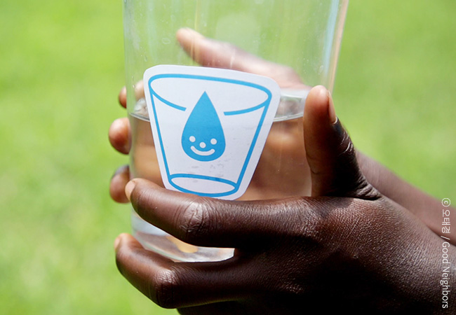 굿워터프로젝트 로고가 붙여 있는 유리컵에 물을 담아 들고 있는 손의 모습